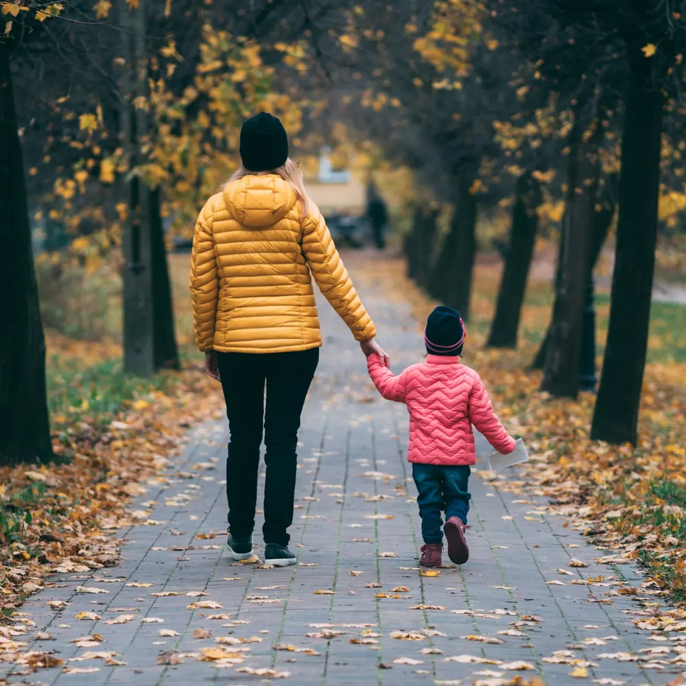 A parent in a yellow jacket walking hand in hand with a small child down a leafy path in the fall